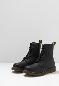 Bottes lacées en cuir noir avec un bout rond, coutures jaunes le long de la semelle et détail de fermeture éclair noire, comportant une tirette jaune.