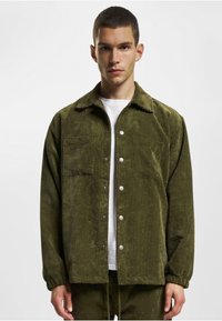 Olive green corduroy jacket with a collar, front pockets, snap buttons, and elastic cuffs, worn over a white t-shirt. Subtle ribbed texture.