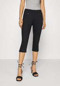 Mujer vestida con pantalones cortos negros de cintura alta con doble botón y sandalias de tacón negro con tiras, de pie contra un fondo liso.