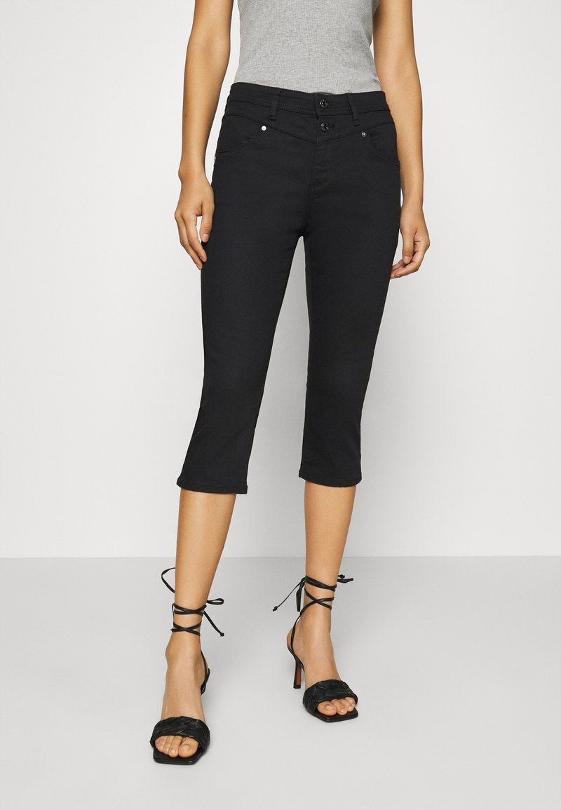 Mujer vestida con pantalones cortos negros de cintura alta con doble botón y sandalias de tacón negro con tiras, de pie contra un fondo liso.