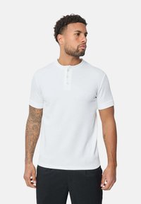 Chemise henley blanche à manches courtes en tissu texturé, avec un col rond et une patte de boutonnage à trois boutons. Design simple, sans motifs.