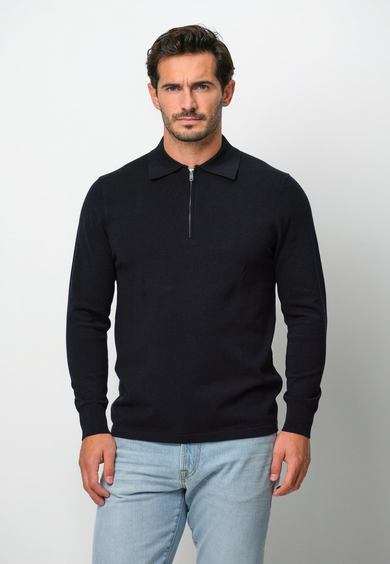 Mörkblå zip-up tröja med spetsig krage och långa ärmar. Tillverkad av mjukt, texturerat tyg. Kombinerad med ljusblå jeans.