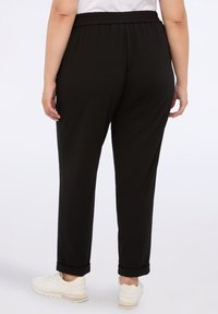 Bakvyn av person som bär svarta elastiska midjebyxor med raka ben och vita sneakers, stående mot en enkel bakgrund.