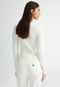 Top bianco a maniche lunghe realizzato in tessuto morbido, con un design aderente e un collo rotondo. Abbinato a jeans bianchi e un piccolo dettaglio logo nero.