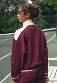 Vīnogu sarkans džemperis ar baltiem akcentiem; liela izmēra piegriezums; ribota aproču un apakšmalas apdare; valkāts kopā ar saskaņotām apakšdaļām; teksturēts audums; sportiska vide.