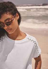 Kvinna med lockigt hår som bär solglasögon i sköldpaddsmönster och en vit skjorta med utskurna spetsdetaljer, stående på en strand med vågor i bakgrunden.