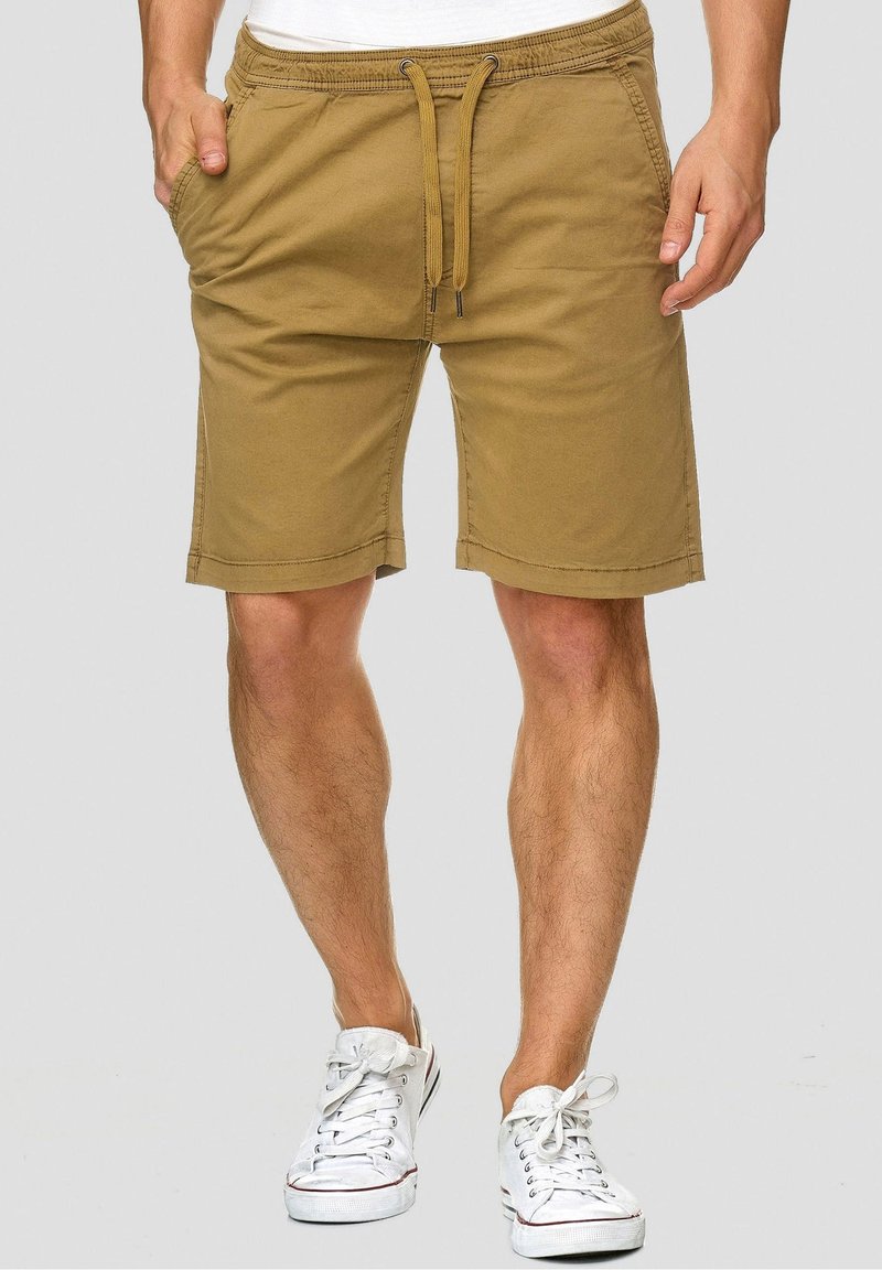 Homme portant un short beige à cordon de serrage et des baskets blanches, debout devant un fond uni gris clair.