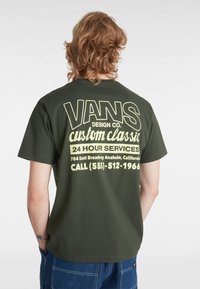Oliven grøn bomulds T-shirt med stor gul tekst på ryggen med "VANS DESIGN CO." og serviceoplysninger. Korte ærmer og afslappet pasform.