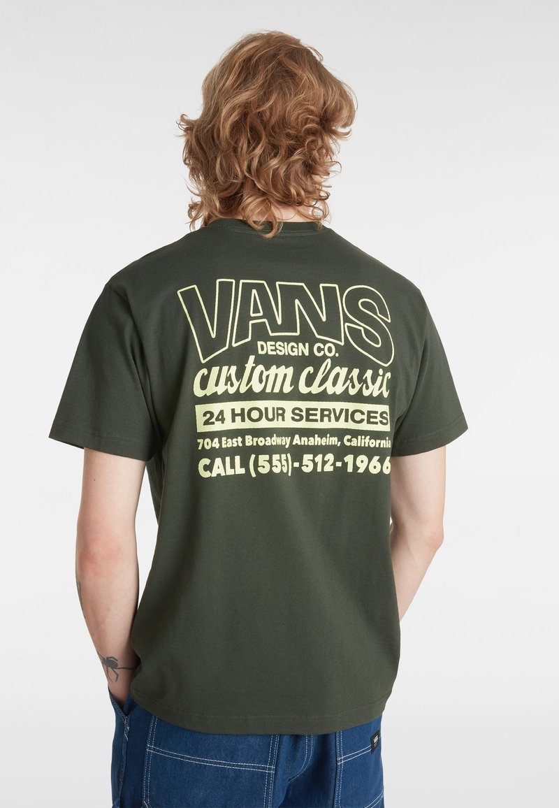 Oliven grøn bomulds T-shirt med stor gul tekst på ryggen med "VANS DESIGN CO." og serviceoplysninger. Korte ærmer og afslappet pasform.
