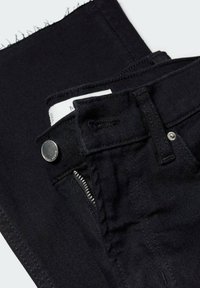 Lähikuva mustista denimpö trousersista, joissa on nähtavad hõbedane nupp, lukku, vöökohad ja tooriku serva detail pükste jala peal.