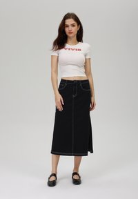 Jupe en denim noir avec des coutures blanches, avec une fente sur le côté, associée à un t-shirt blanc ajusté portant le mot "VIVID". Chaussures noires plates.