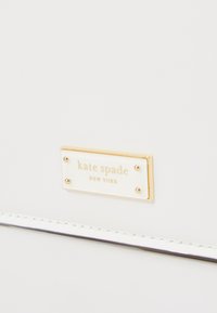 Bijela torbica s zlatnim pravokutnim oznakom na kojoj je ugravirano "kate spade NEW YORK", s glatkom kožnom teksturom i detaljima šivanja.