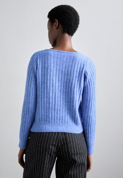 Pulover ribbed de culoare bleu deschis, cu o decolteu larg și mâneci lungi, fabricat dintr-un amestec moale de lână. Asortat cu pantaloni în dungi întunecate.