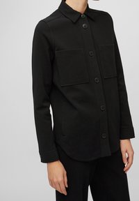 Černá košile s knoflíky z hladkého materiálu, která má dvě náprsní kapsy, klasický límec a dlouhé rukávy s knoflíkovými manžetami.