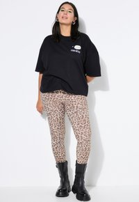 Žena v černém volném tričku s potiskem, béžové legíny s leopardím vzorem, černé robustní boty, stojí před bílým pozadím.