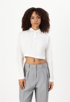 Bílé krácené button-up košile s dlouhými rukávy. Vyrobená z lehkého materiálu, s klasickým límcem a hladkou texturou. V kombinaci se šedými skládacími kalhotami.