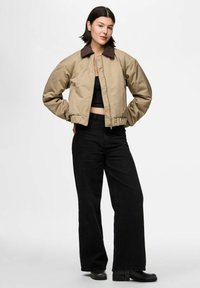 Sieviete, kas ģērbjas bēšā jakā ar brūnu apkakli, melnā kroptopā, platās melnās biksēs un melnās zābakos, stāv ar rokām aiz muguras.