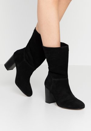 Jambes portant des bottes mi-mollet en daim noir avec des talons blocs empilés sur une surface gris clair.