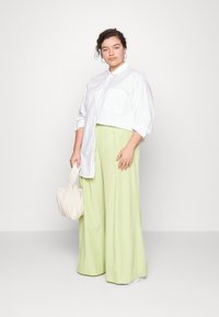 Camisa oversized branca, calças largas verdes claras e uma bolsa texturizada bege. Modelo em pé com um fundo neutro, com detalhes mínimos.