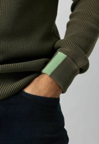 Olive green knitted sweater featuring ribbed texture and a light green accent on the cuff, paired with dark blue denim jeans.
