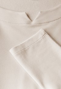 Beige kangas, joka esittelee sileää pintaa ja V-pääntien muotoilua, näkyvillä ompeleilla. Näytetään myös lisäosa, joka on taitettu samasta materiaalista.