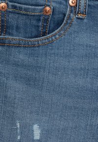 Tessuto di denim blu con una zona logora, caratterizzato da dettagli in rame alla vita e cuciture gialle lungo le tasche e le cuciture.