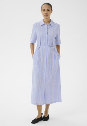 Femme debout portant une robe midi rayée bleu clair à boutons, manches courtes, et des chaussures noires à enfiler, sur un fond blanc uni.