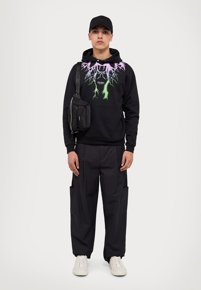 Felpa nera con grafica di fulmini viola, pantaloni neri con tasche laterali, sneakers bianche e una piccola borsa a spalla nera, indossata con un cappellino.