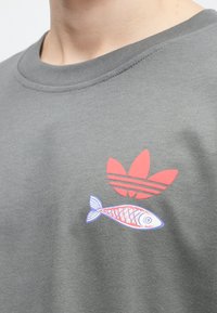 Pelēks kokvilnas sporta krekls ar sarkano Adidas logo un stilizētu zivs grafiku zilā un rozā krāsā, ar apaļu apkakli.