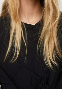Černý prošívaný oděv s výstřihem do V a zapínáním na knoflíky, s dlouhými rovnými blond vlasy částečně viditelnými v záběru.