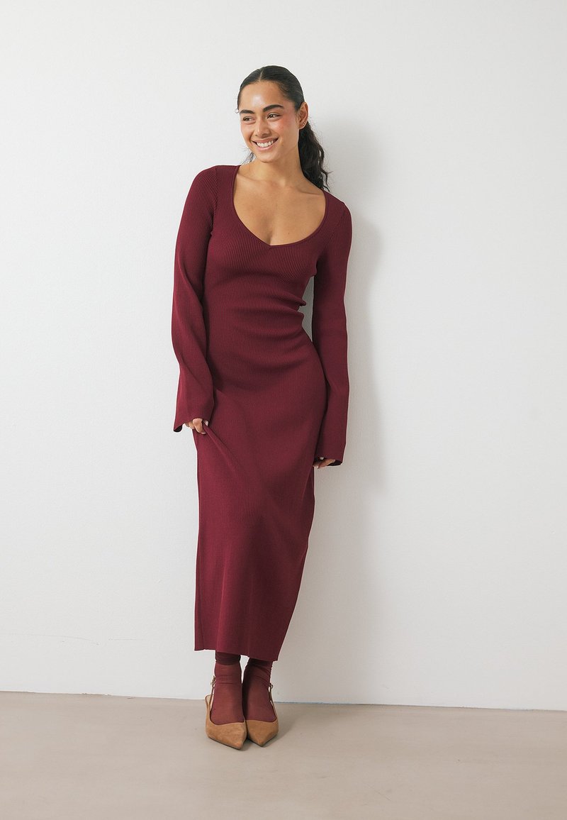 Robe en tricot côtelé bordeaux avec un décolleté plongeant, des manches longues et une silhouette ajustée, associée à des bottines assorties.