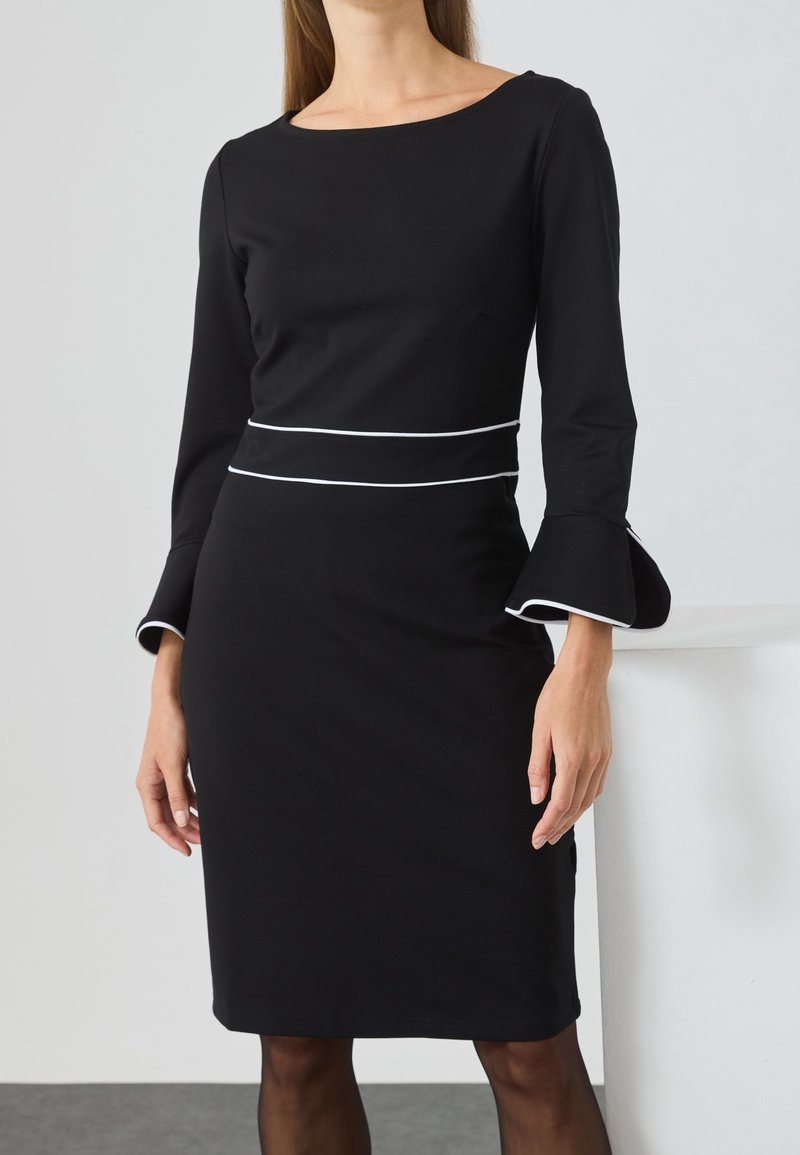 Femme portant une robe noire jusqu'aux genoux avec une bordure blanche à la taille et des manches cloche, debout devant un fond clair uni.