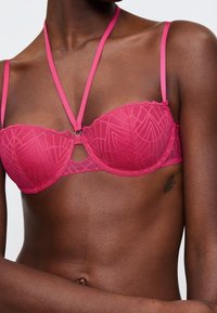 Růžová krajková podprsenka s geometrickým vzorem, s kosticemi, zvlněnými okraji a tenkými nastavitelnými ramínky s dekorativním předním akcentem.