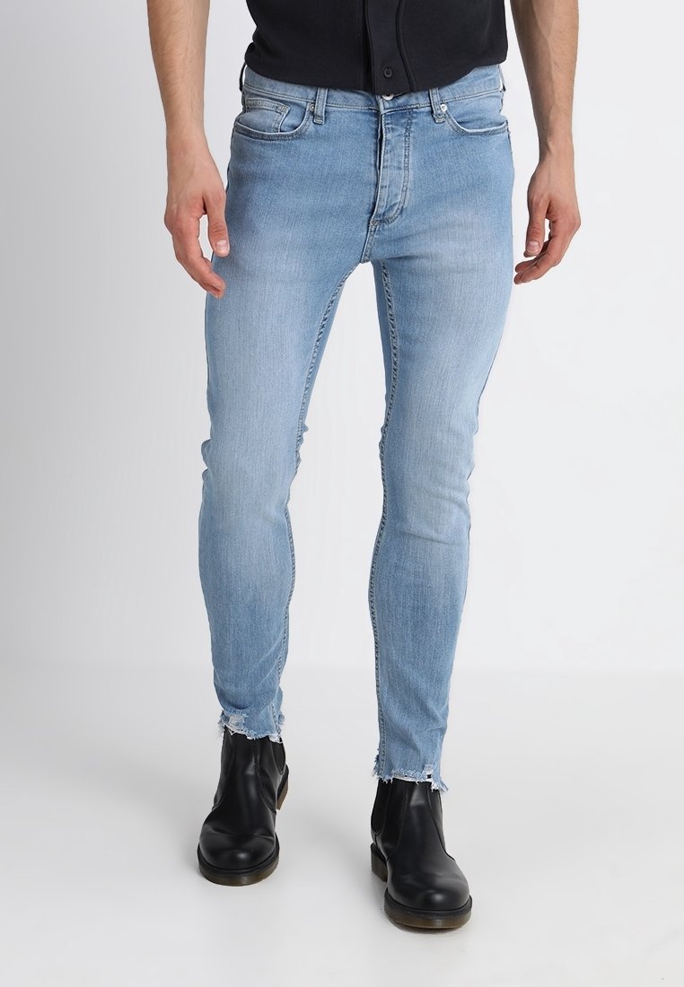 Homme portant un jean skinny bleu clair avec des ourlets bruts et des bottines noires, debout devant un fond blanc uni.