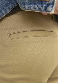 Tessuto khaki con una trama texturizzata, caratterizzato da una piccola tasca cucita sul retro. Parte di un indumento inferiore di una persona è parzialmente visibile.