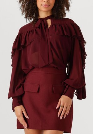 Vrouw in een bordeauxrode blouse met ruches en een strik om de nek, gecombineerd met een bijpassende high-waisted rok met voorklepzakken.