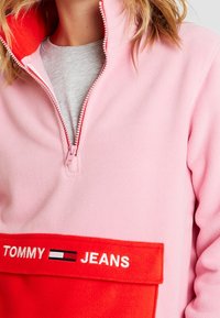 Różowy polarowy pulower z czerwoną kieszenią z przodu. Posiada kołnierz z pół-zamkiem oraz białe haftowane napisy "TOMMY JEANS". Miękka tekstura.