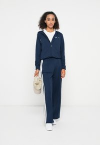 Survêtement bleu marine avec des bandes blanches sur les côtés, veste à capuche zippée, poignets élastiques et pantalon ample assorti. Tenant une casquette beige.