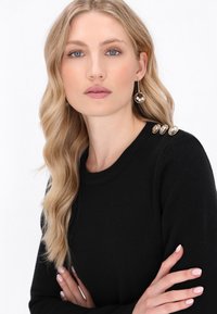 Fekete pulóver kerek nyakkivágással, bal vállán négy díszgombbal; a modellen hullámos haj és kör alakú fülbevalók találhatók.
