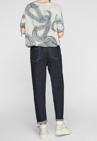 Maglietta a maniche corte con motivo in tessuto leggero e disegni astratti blu; abbinata a pantaloni in denim scuro e sneakers di colore chiaro.