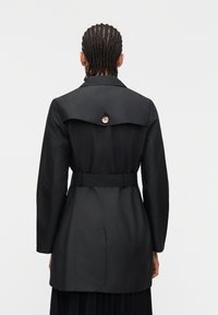 Άτομο με κοτσίδα, που φοράει μαύρο παλτό με ζώνη, εμφανιζόμενο από πίσω πάνω σε απλό λευκό φόντο.