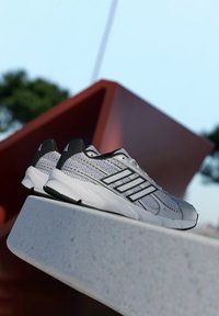 Ľahké atletické topánky s očkovou hornou časťou v sivej farbe s čiernymi akcentmi. Dizajn obsahuje tri biele pruhy po bokoch a mäkkú podrážku.