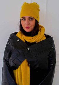 Veste en simili cuir noire avec des boutons-pression, superposée à une écharpe jaune vif et un bonnet assorti, créant un contraste de couleurs audacieux.
