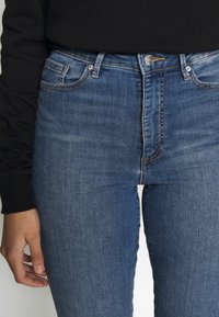 Jeans de mezclilla azul de talle alto con ajuste Slim, que cuentan con cinco bolsillos, costuras claras y un cierre de botón de metal en la cintura.