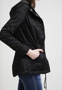 Czarna kurtka z kapturem, ze sznurkiem w talii, przednim zamkiem błyskawicznym i bocznymi kieszeniami. Wykonana z gładkiego materiału o luźnym kroju.