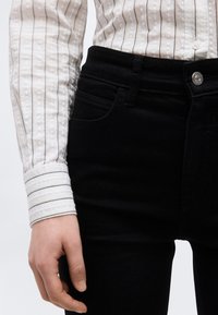 Oseba, ki nosi belo majico z dolgimi rokavi s črnimi črtami, zataknjeno v visoko pasne črne kavbojke s srebrnim gumbom.