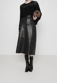 Jupe midi en cuir noir à taille haute et texture lisse. Associée à un pull en maille noir à franges et des bottes noires à bout pointu.