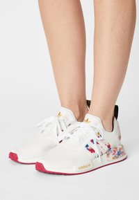 White athletic shoes with colorful floral patterns, pink rubber sole, and white laces. Features a gold logo and black heel tab.
