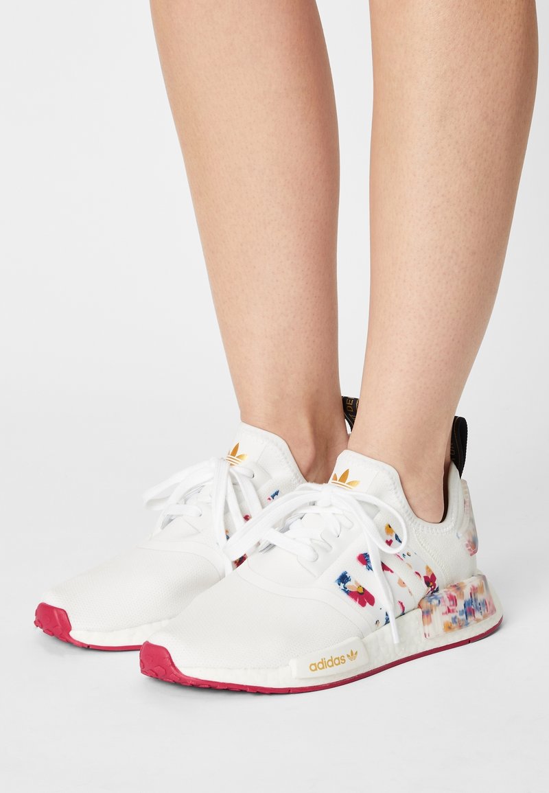 White athletic shoes with colorful floral patterns, pink rubber sole, and white laces. Features a gold logo and black heel tab.