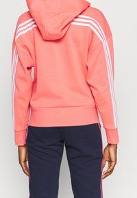 Felpa corallo con design a tre strisce bianche lungo le maniche, orlo a coste e dettagli sui polsini. Indossata con pantaloni blu scuro. Tessuto morbido.